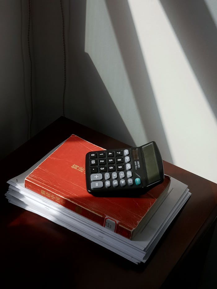 A calculator rests on a red book and stack of papers, basking in sunlight.